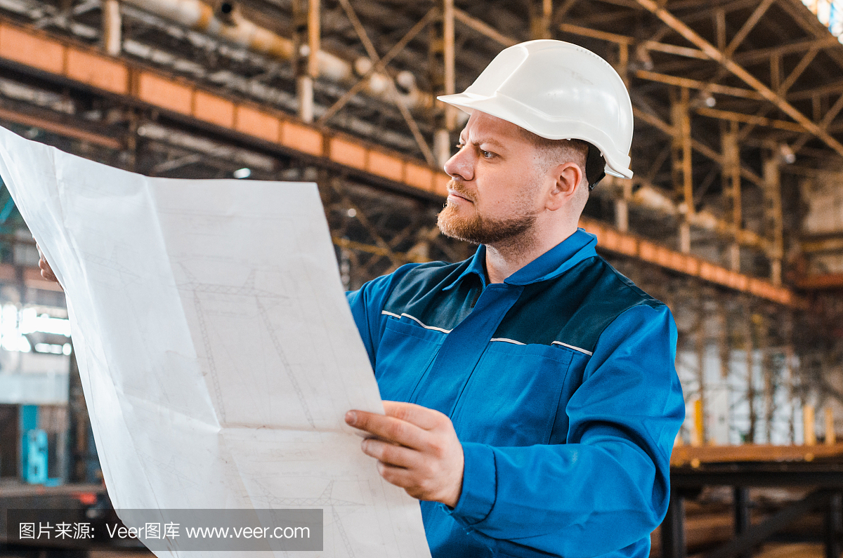 一個穿著工作服、戴著白色頭盔的工人正在檢查一個金屬工廠背景上的圖紙。建筑工人身體很好。男工程師